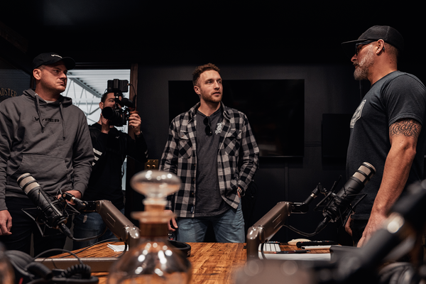four guys in a studio standing around a table in discussion. there are bottles of whiskey and microphones on the table