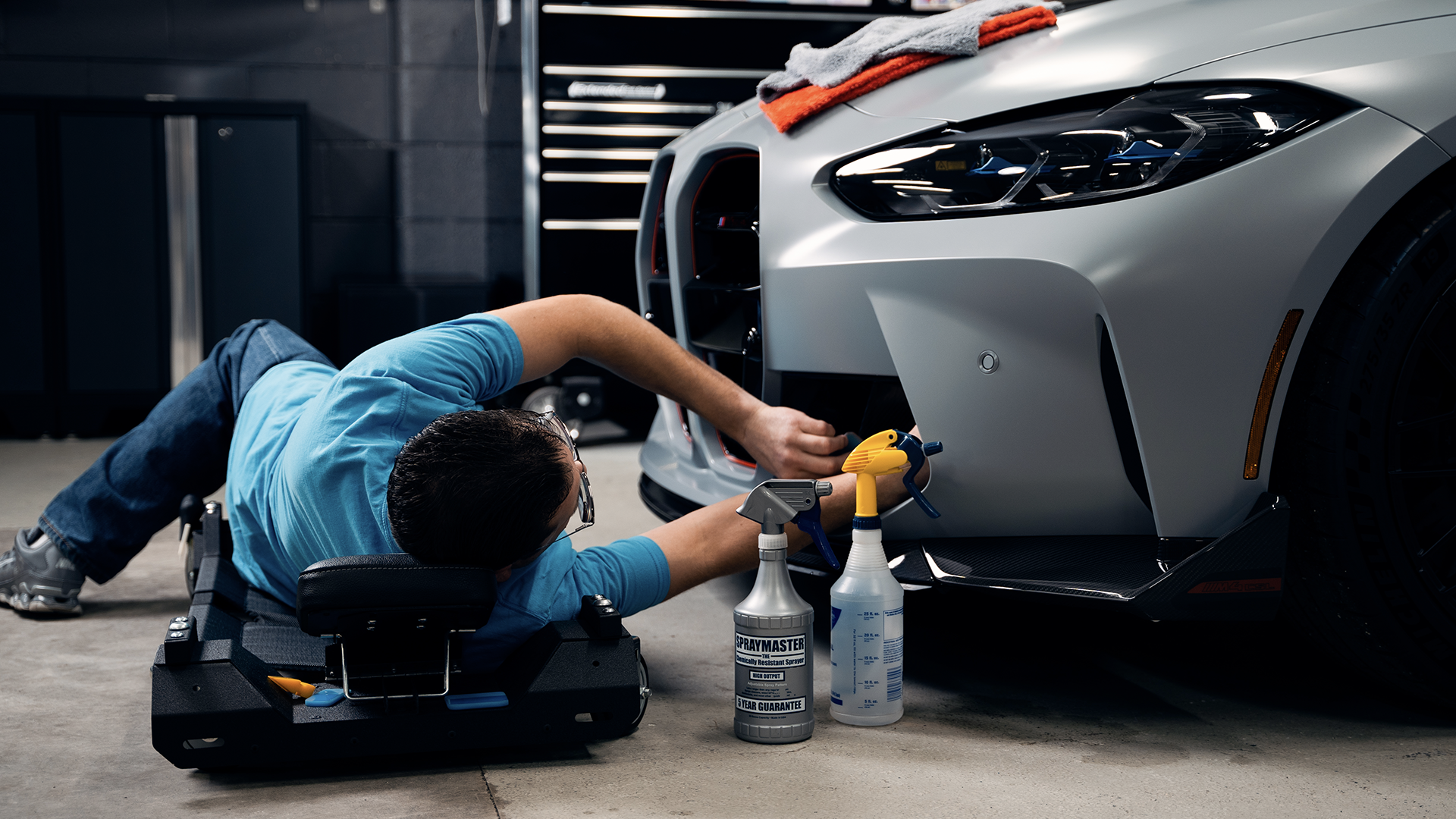 Man in light blue shirt laying down on creeper working on gray car