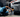 Man in light blue shirt laying down on creeper working on gray car