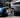 Man in light blue shirt laying down on creeper working on gray car