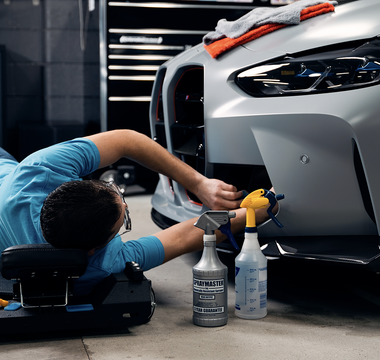 Man in light blue shirt laying down on creeper working on gray car