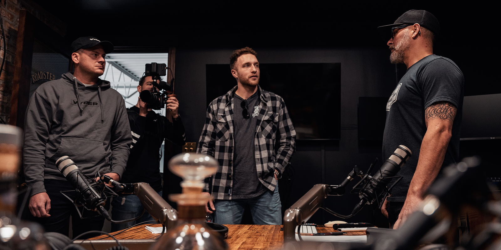 four guys in a studio standing around a table in discussion. there are bottles of whiskey and microphones on the table