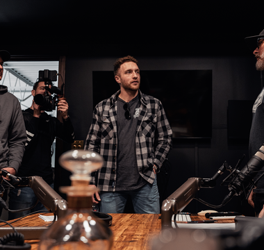 four guys in a studio standing around a table in discussion. there are bottles of whiskey and microphones on the table