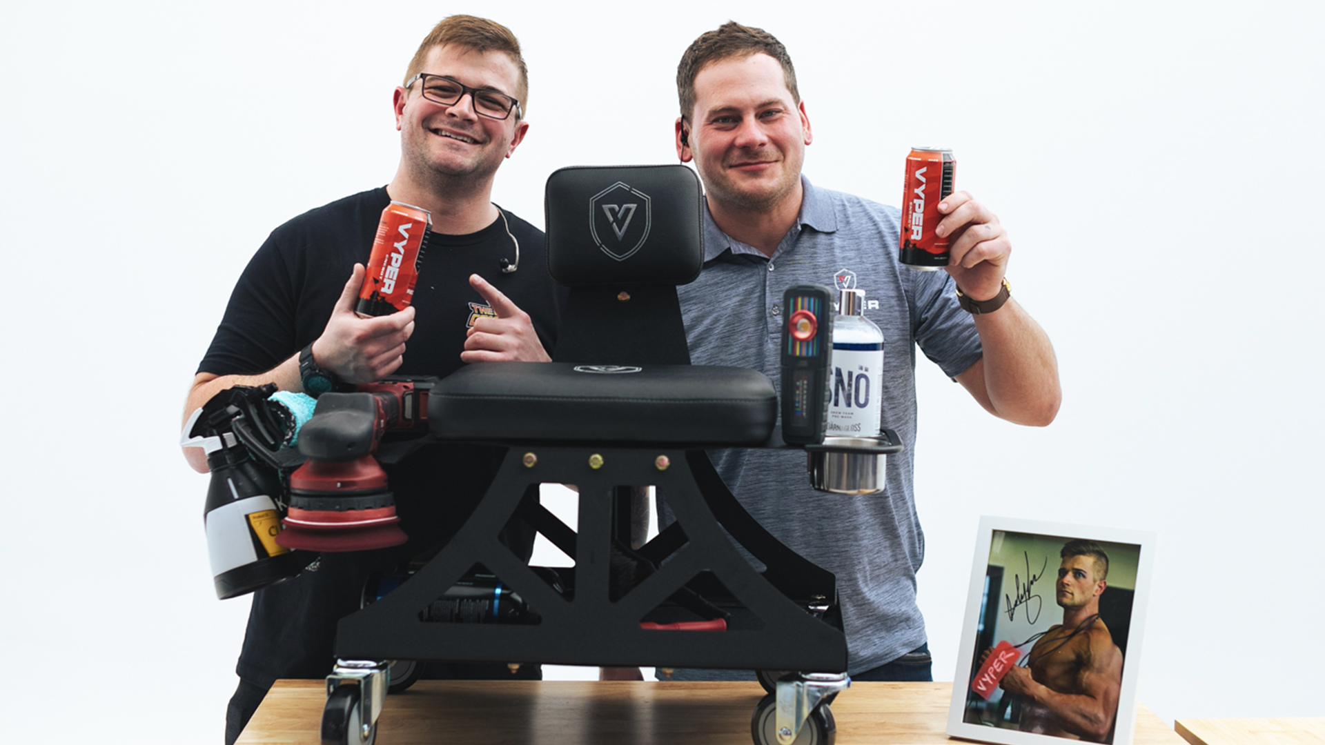 Two guys behind an industrial shop chair on wheels sitting on a table. Both are holding red energy drink cans and smiling