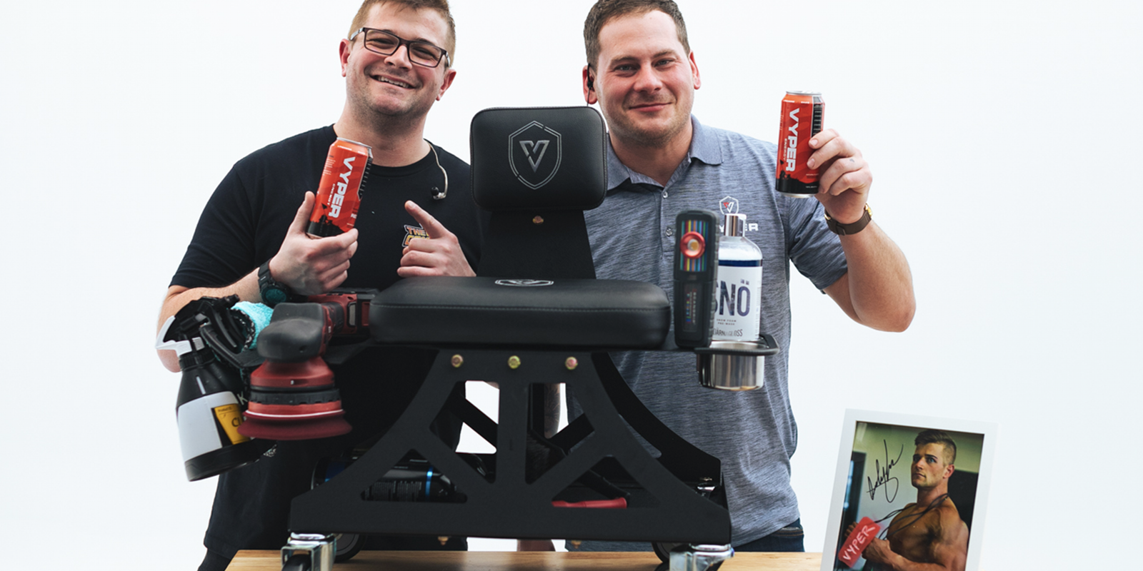 Two guys behind an industrial shop chair on wheels sitting on a table. Both are holding red energy drink cans and smiling