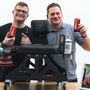 Two guys behind an industrial shop chair on wheels sitting on a table. Both are holding red energy drink cans and smiling